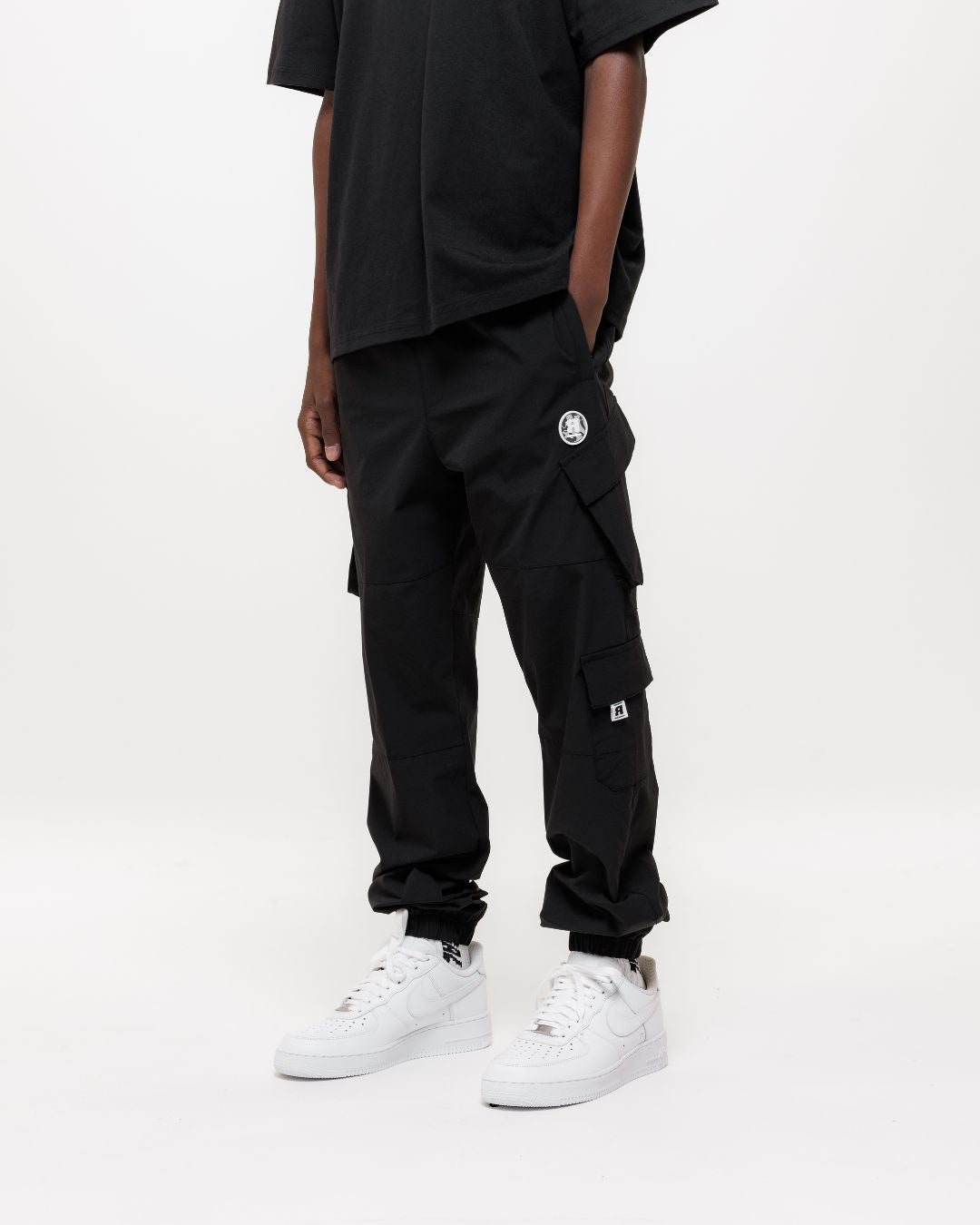 boy wearing next gen black junior cargo pants and tee with Rascal camo logo designs, hand in pockets stood in front of a white background