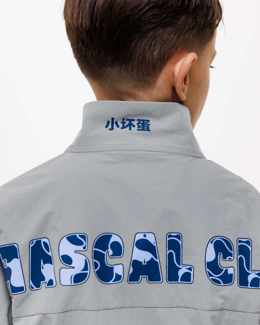 boy wearing next gen grey junior shell jacket with Rascal camo text design across the back and Chinese symbols on the collar