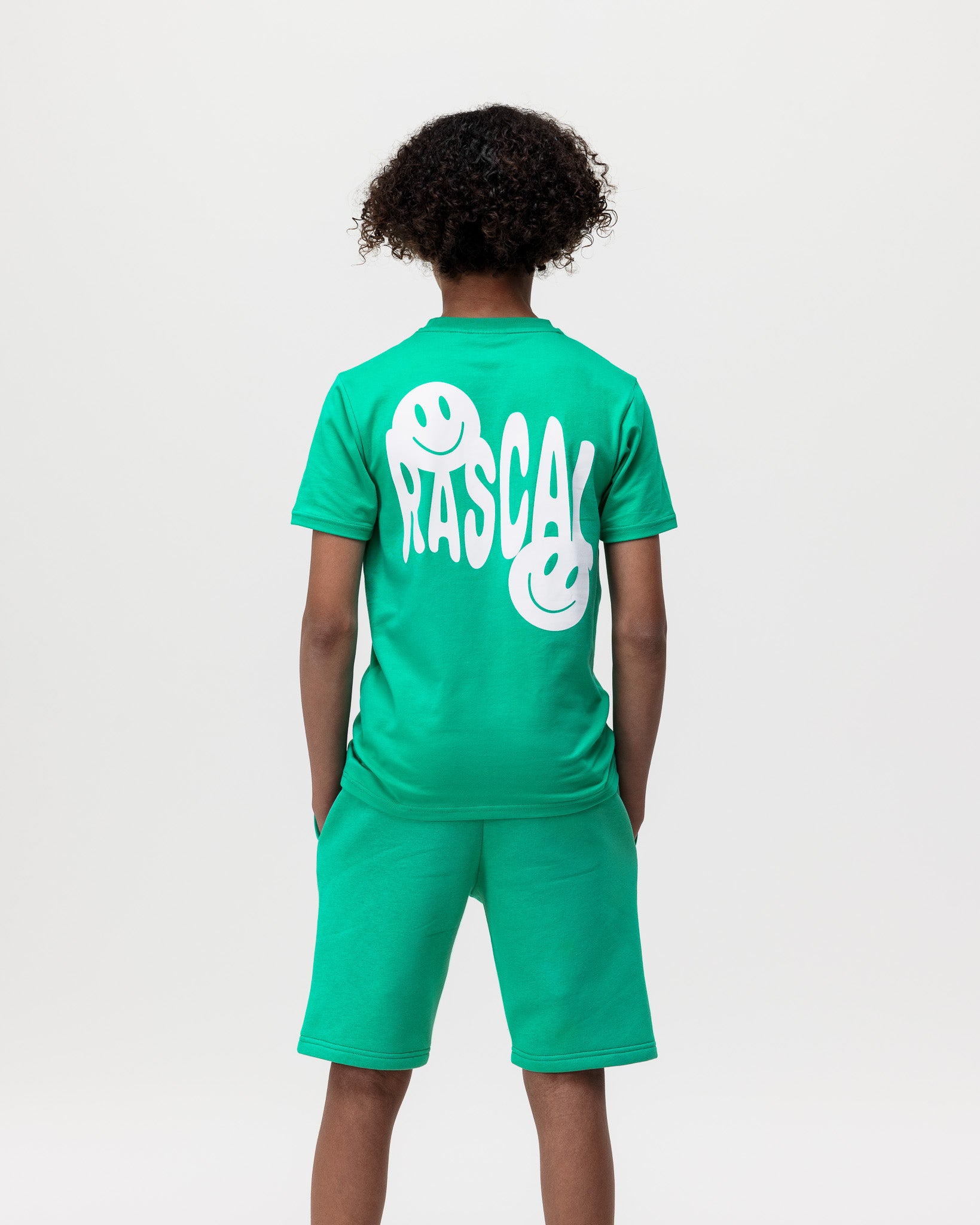 boy standing with hands in his pockets wearing Rascal green shorts and green smiley face tshirt, showing the back of tshirt with white Rascal and smiley face graphics 