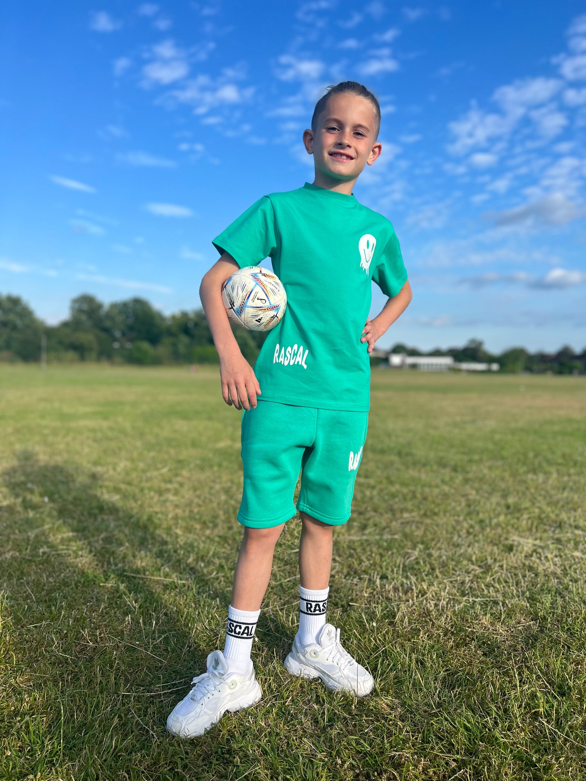 Roman Wingrove standing with football under his arm wearing green Rascal shorts and green smiley face tshirt with white Rascal and smiley face graphics