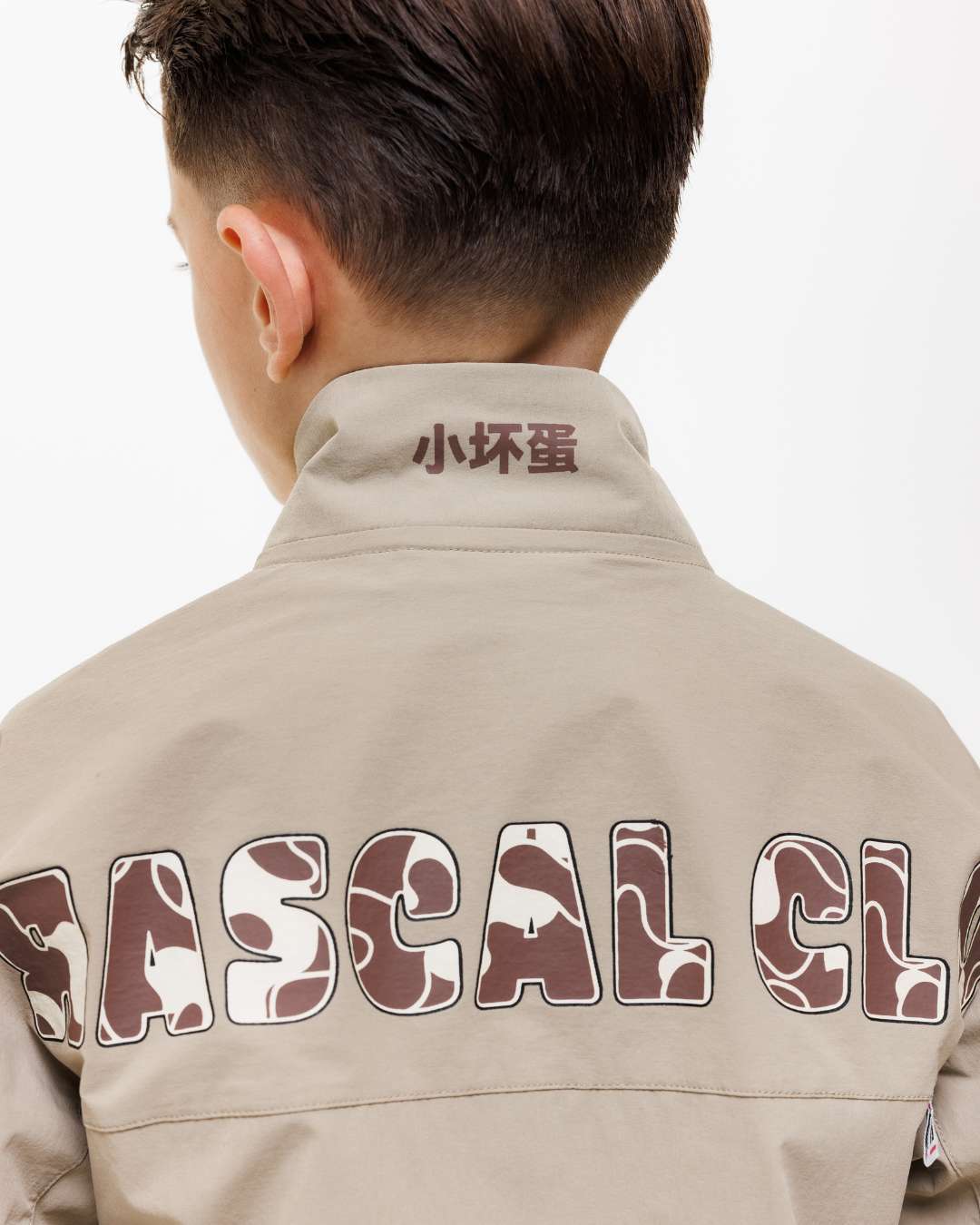 Boy wearing a Next Gen Junior khaki jacket showing Rascal Camo text printed on the back and Chinese symbols on the collar
