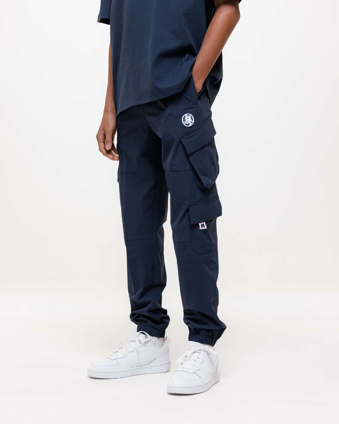 Boy wearing Next Gen junior navy blue cargo pants including Rascal design elements with hand in pocket and tee stood in front of a white background 