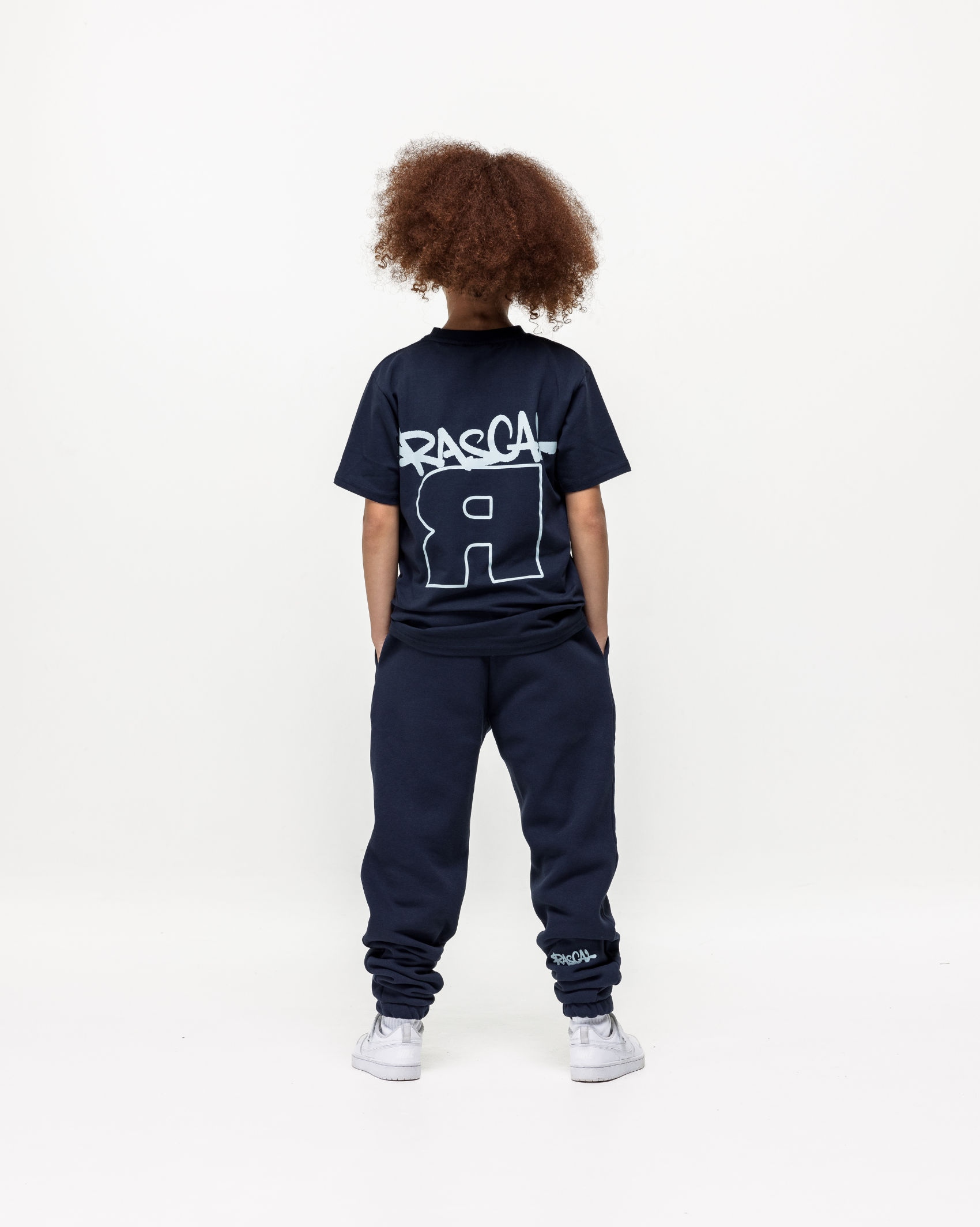 Girl model wearing Rascal Flip Navy Joggers and matching tee, showing the back of both items stood with hands in pocket