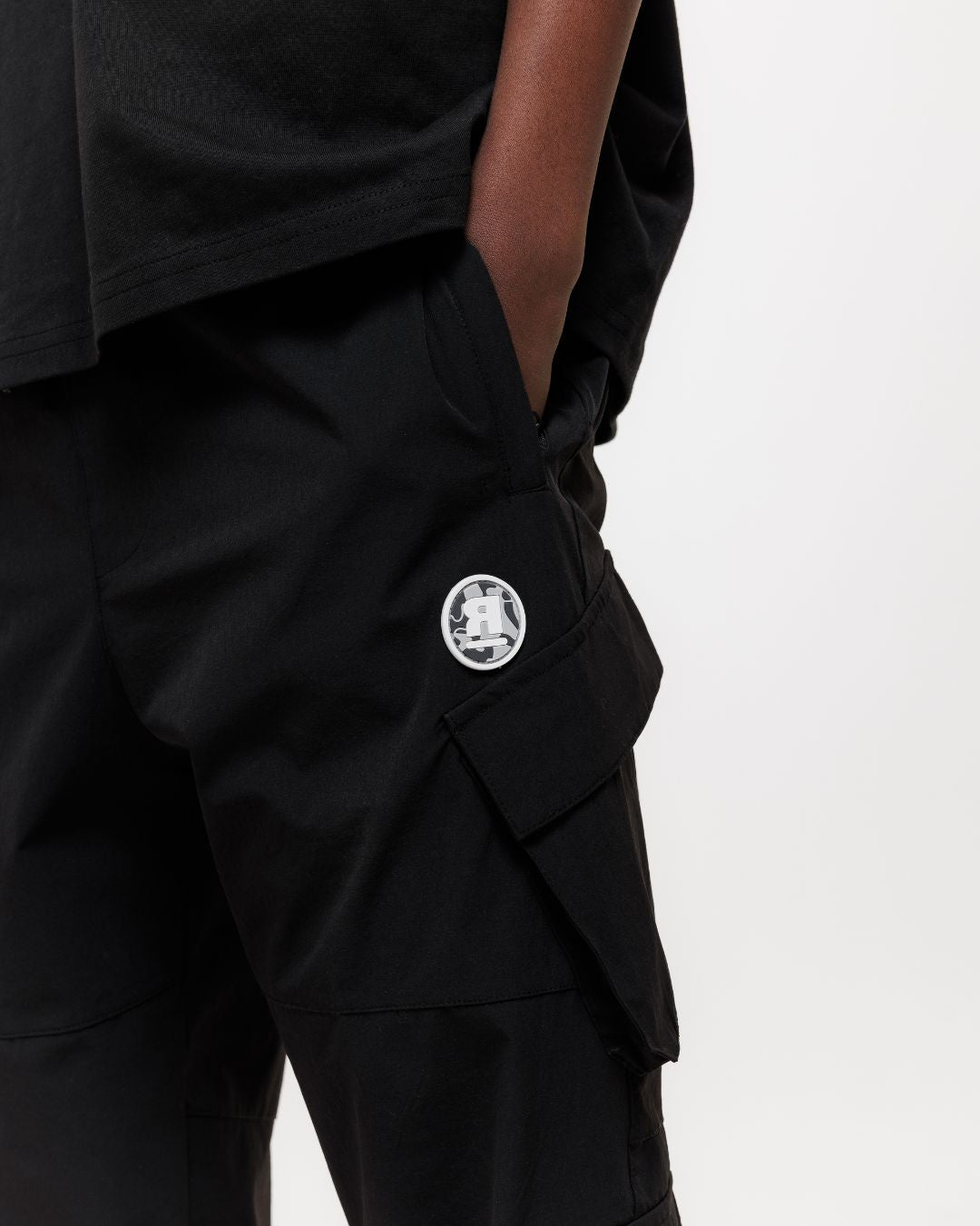 close up of next gen black junior cargo pants with Rascal camo logo designs, hand in pocket stood in front of a white background