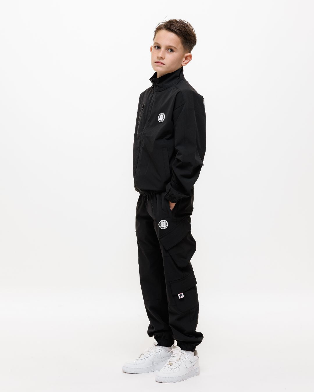 boy wearing next gen black junior shell jacket and cargo pants with Rascal camo logo designs, hands in pockets stood in front of a white background