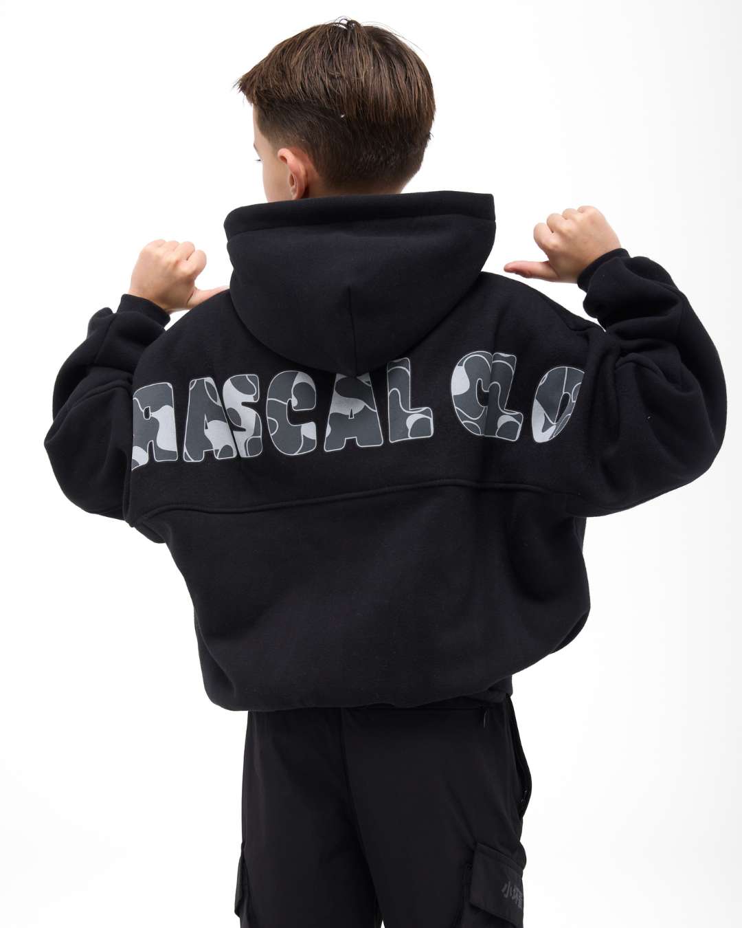 boy wearing next gen black junior oversized hoodie lifting arms and pointing at Rascal camo text design stood in front of a white background