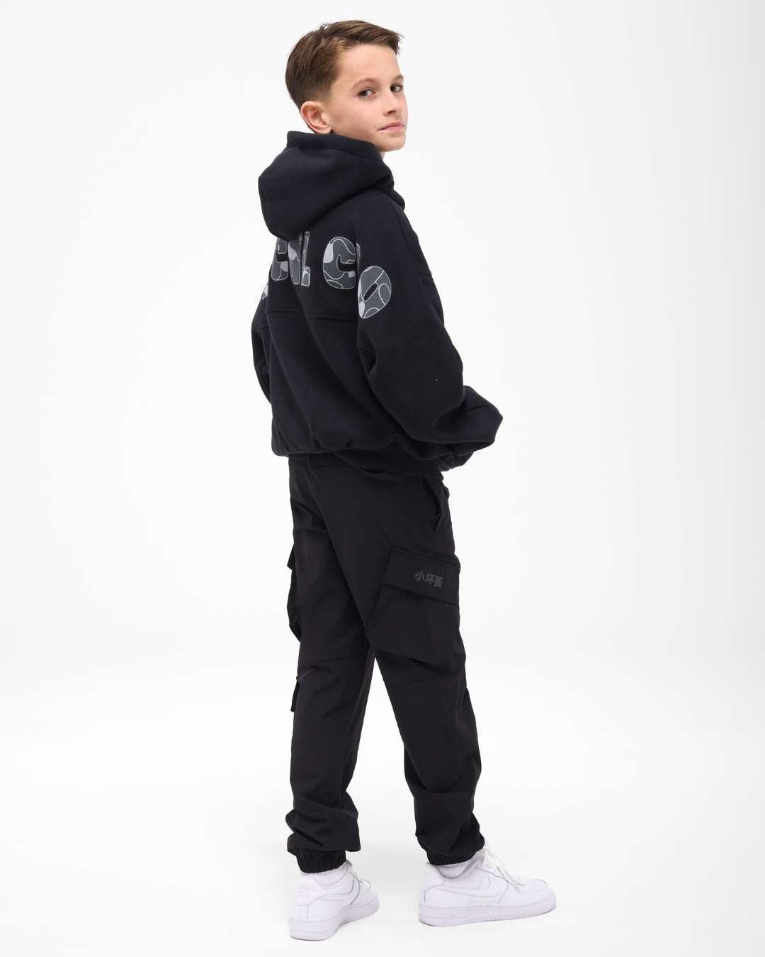 boy wearing next gen black junior oversized hoodie with Rascal camo text design and cargos, stood in front of a white background looking over his shoulder