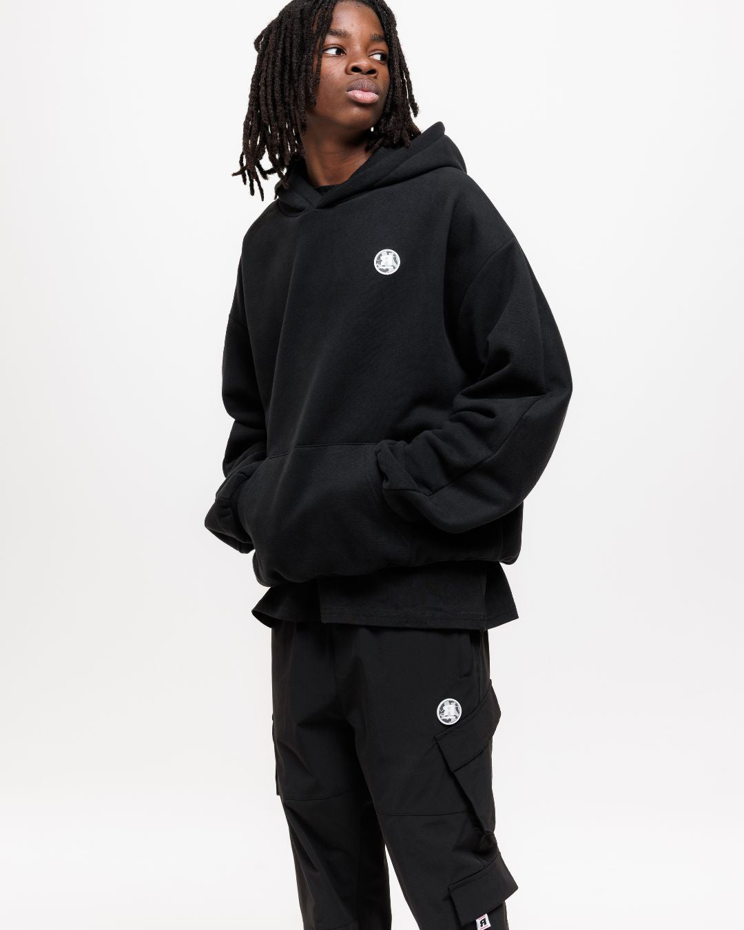 boy wearing next gen black junior oversized hoodie and cargo pants with Rascal camo logo design stood in front of a white background with hands in kangaroo pocket 