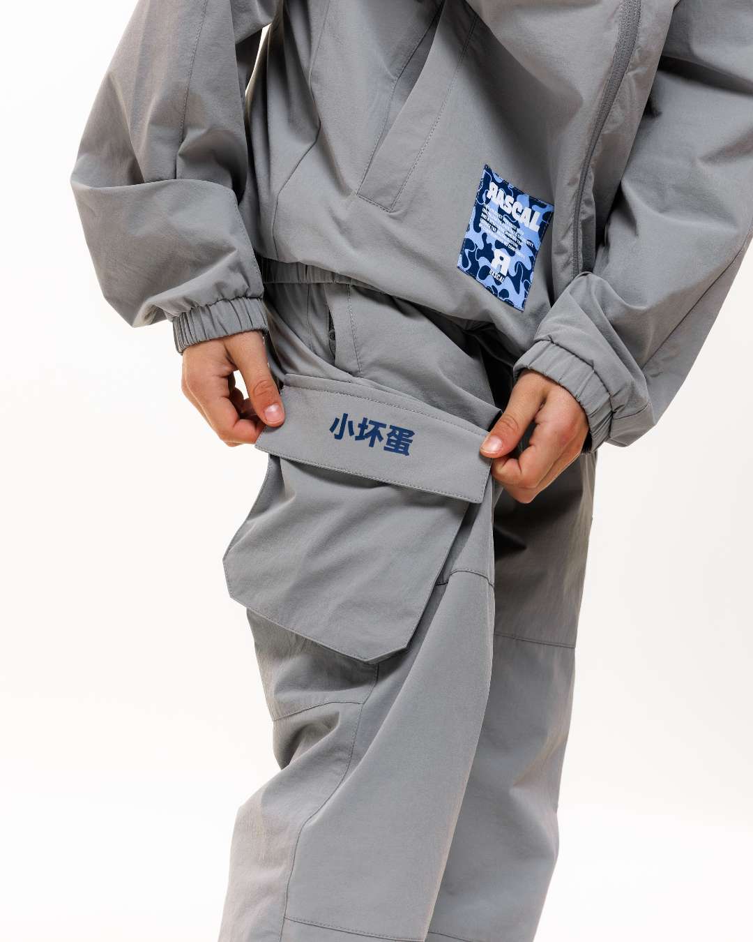 boy wearing next gen grey junior cargo pants and jacket with Rascal camo logo design, stood in front of a white background holding a cargo pant pocket with Chinese symbols design
