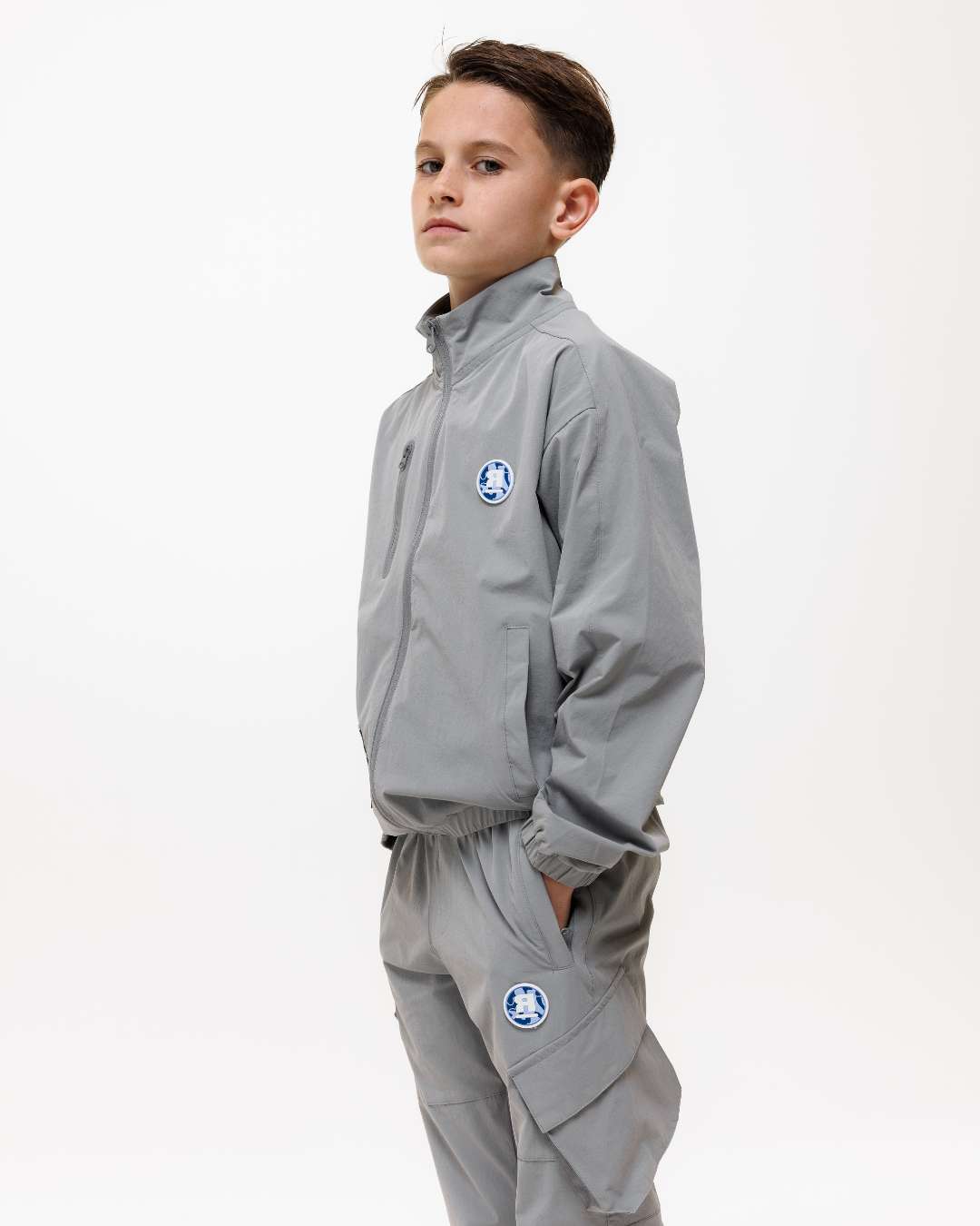 boy wearing next gen grey junior shell jacket and cargo pants with Rascal camo logo design across the back, stood in front of a white background and hands in pockets