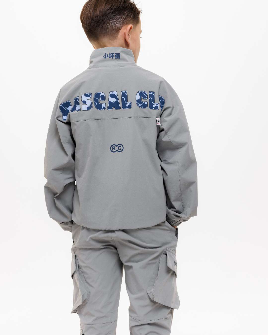 boy wearing next gen grey junior shell jacket and cargo pants with Rascal camo text design across the back, stood in front of a white background and hands in pockets