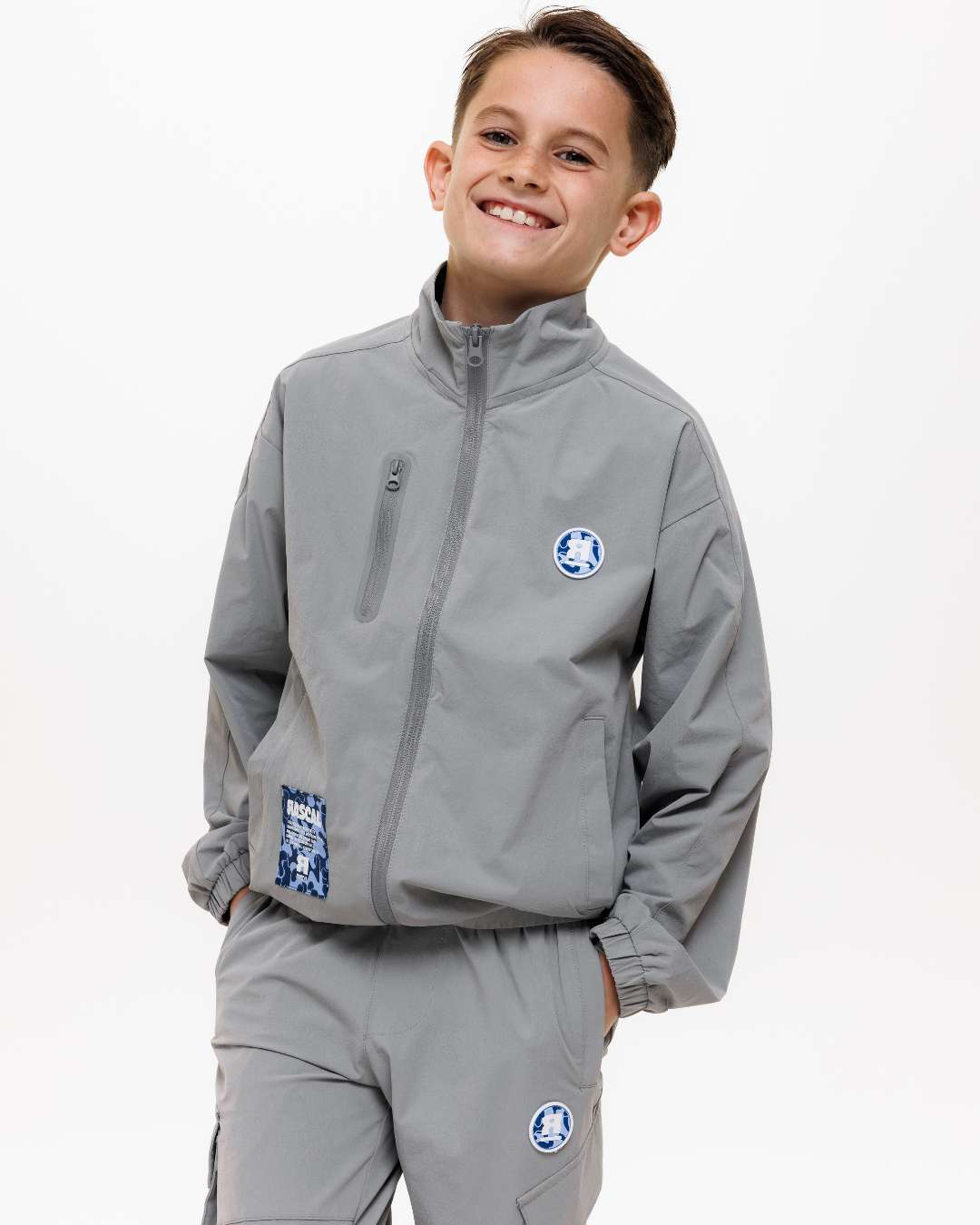boy wearing next gen grey junior shell jacket and cargo pants with Rascal camo logo design, stood in front of a white background and hands in pockets
