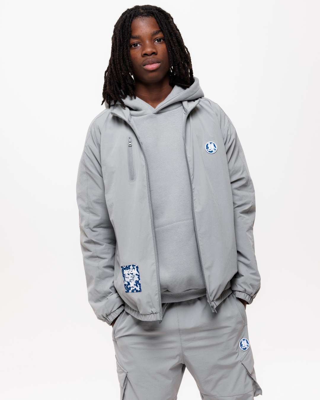 boy wearing next gen grey junior shell jacket, hoodie and cargo pants with Rascal camo logo designs, stood in front of a white background and hands in pockets