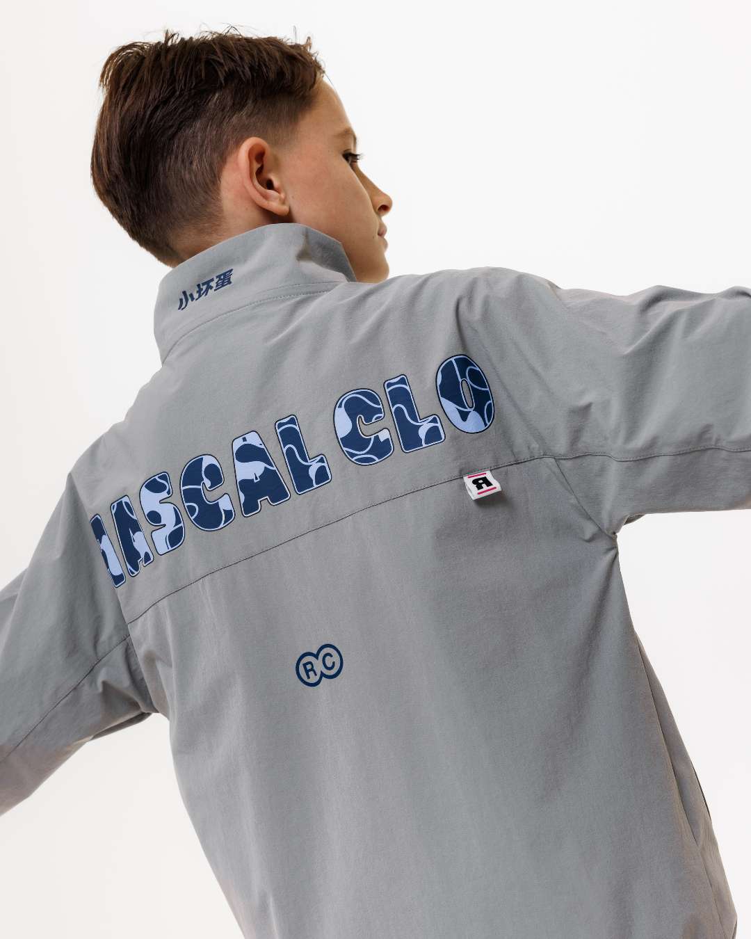 boy wearing next gen grey junior shell jacket with Rascal camo text design across the back, stood in front of a white background and arms lifted to his side