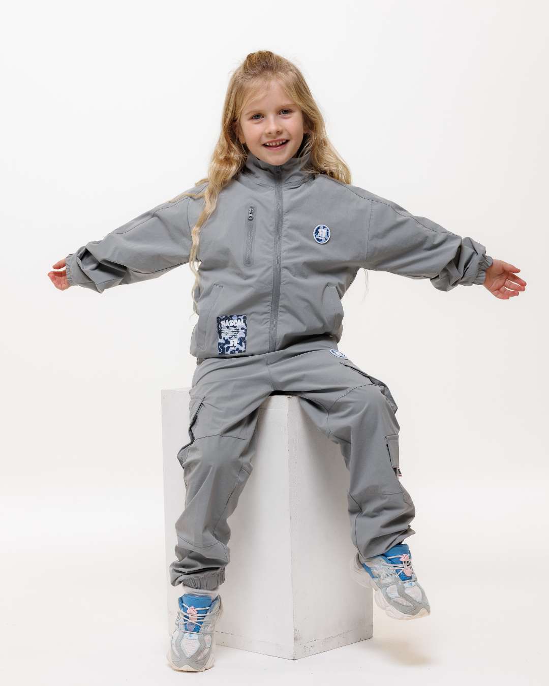 girl wearing next gen grey junior shell jacket and cargo pants with Rascal camo logo design sat on a white block and arms out to her side