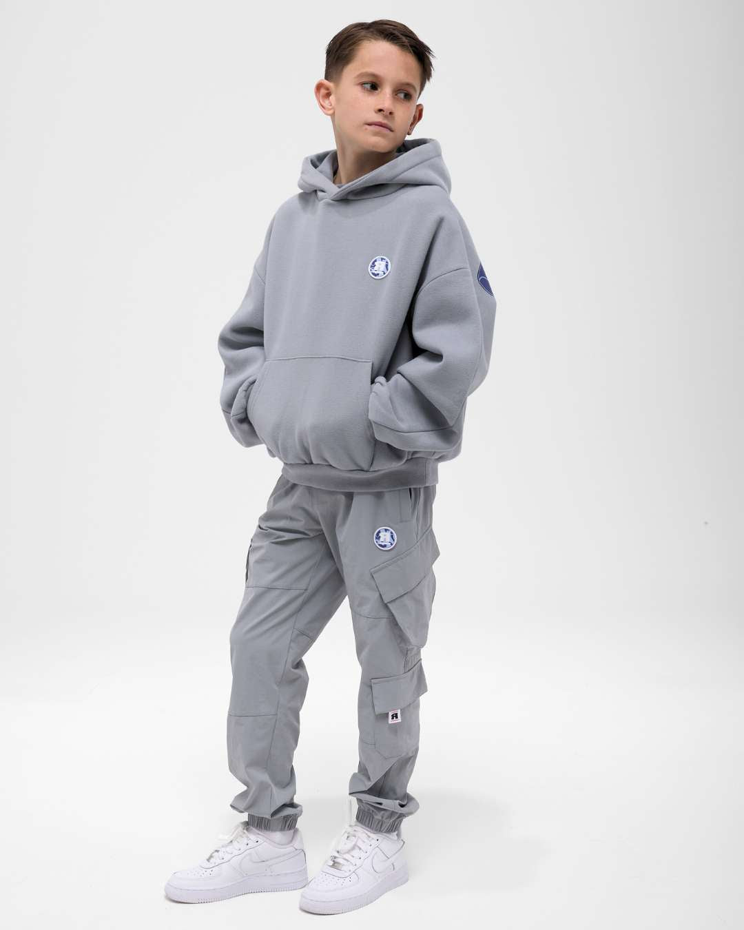 boy wearing next gen junior grey oversized hoodie and cargo pants with Rascal camo logo text designs, stood in front of a white background and hands in kangaroo pocket