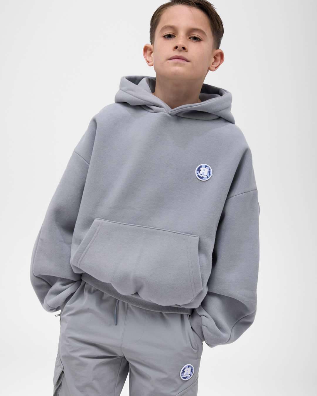 boy wearing next gen junior grey oversized hoodie and cargo pants with Rascal camo logo design, stood in front of a white background