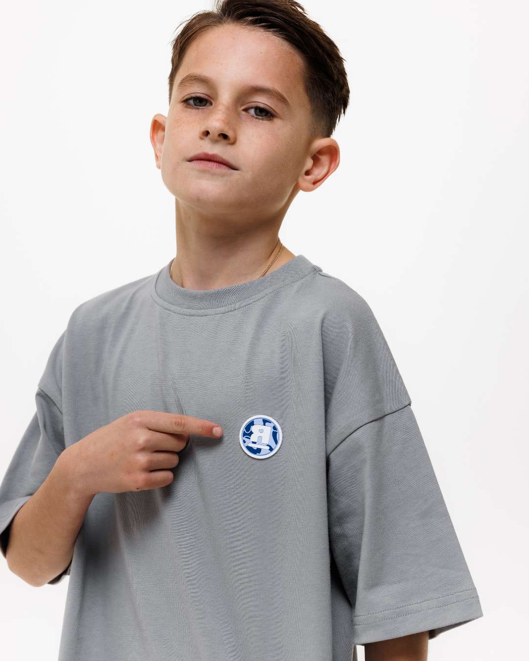 boy wearing next gen junior grey oversized tee pointing at Rascal camo logo design in front of a white background