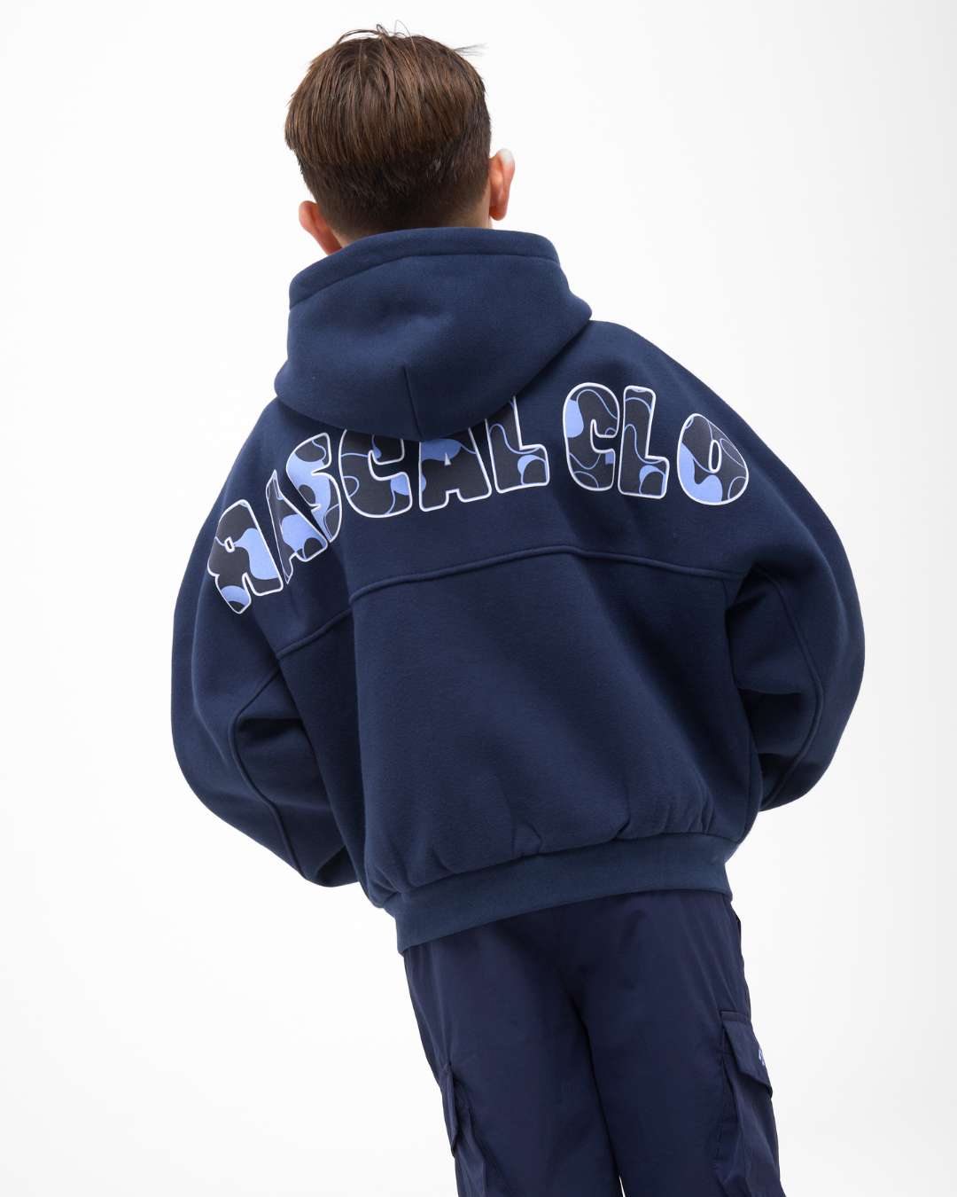 boy wearing next gen navy blue junior oversized hoodie with Rascal camo text design with hands sat in kangaroo pocket on the back against a white background