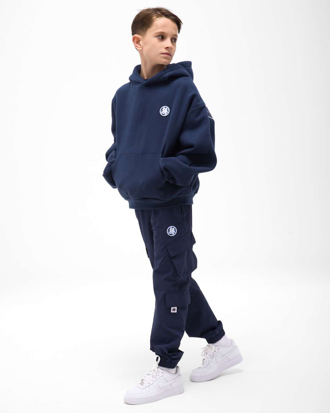 boy wearing next gen navy blue junior oversized hoodie and cargo pants with Rascal camo logo designs, hands inside kangaroo pocket stood in front of a white background