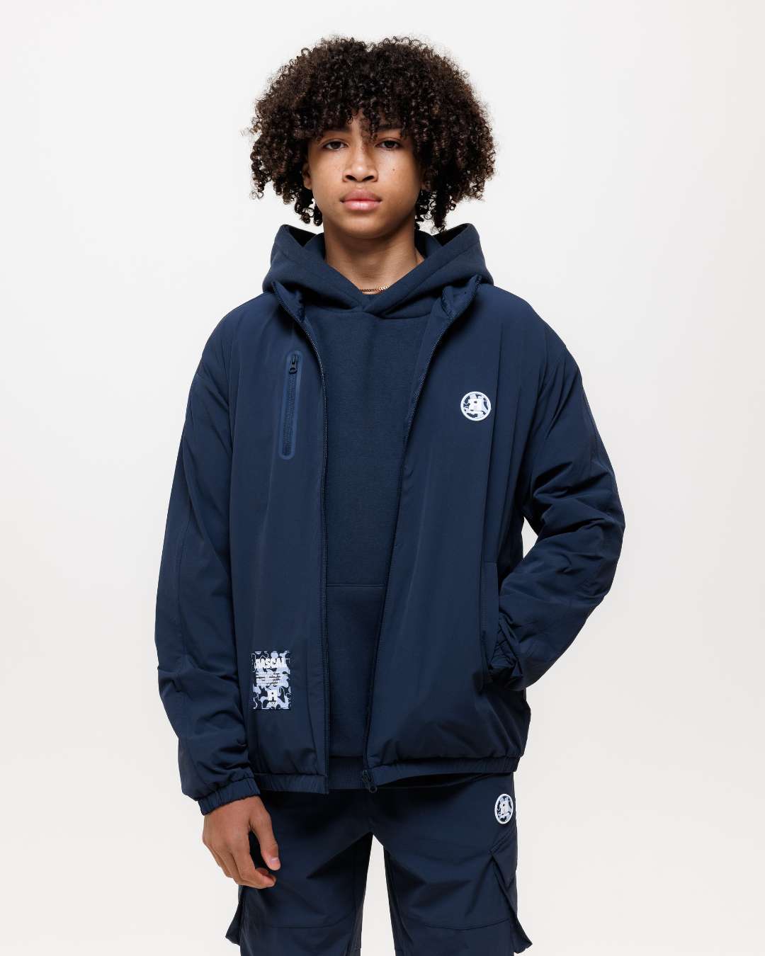 Boy wearing next gen junior navy blue jacket, hoodie and cargos with Rascal designs stood in front of a white background