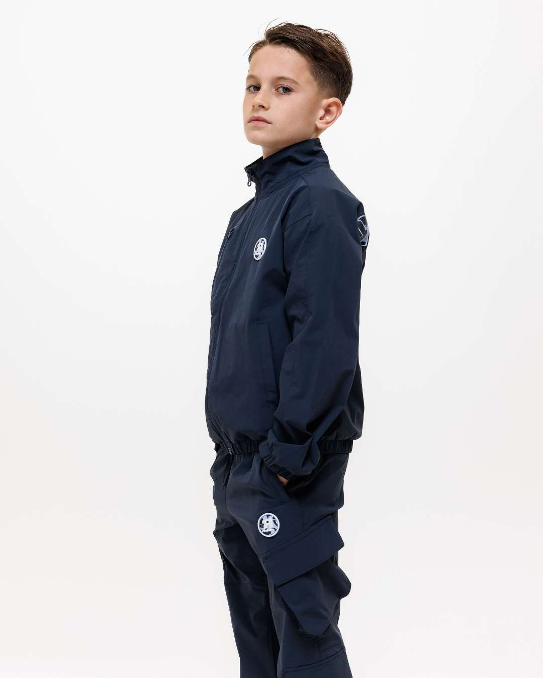 side view of boy wearing next gen junior navy blue jacket and cargo pants tracksuit with Rascal designs and hands in pockets 